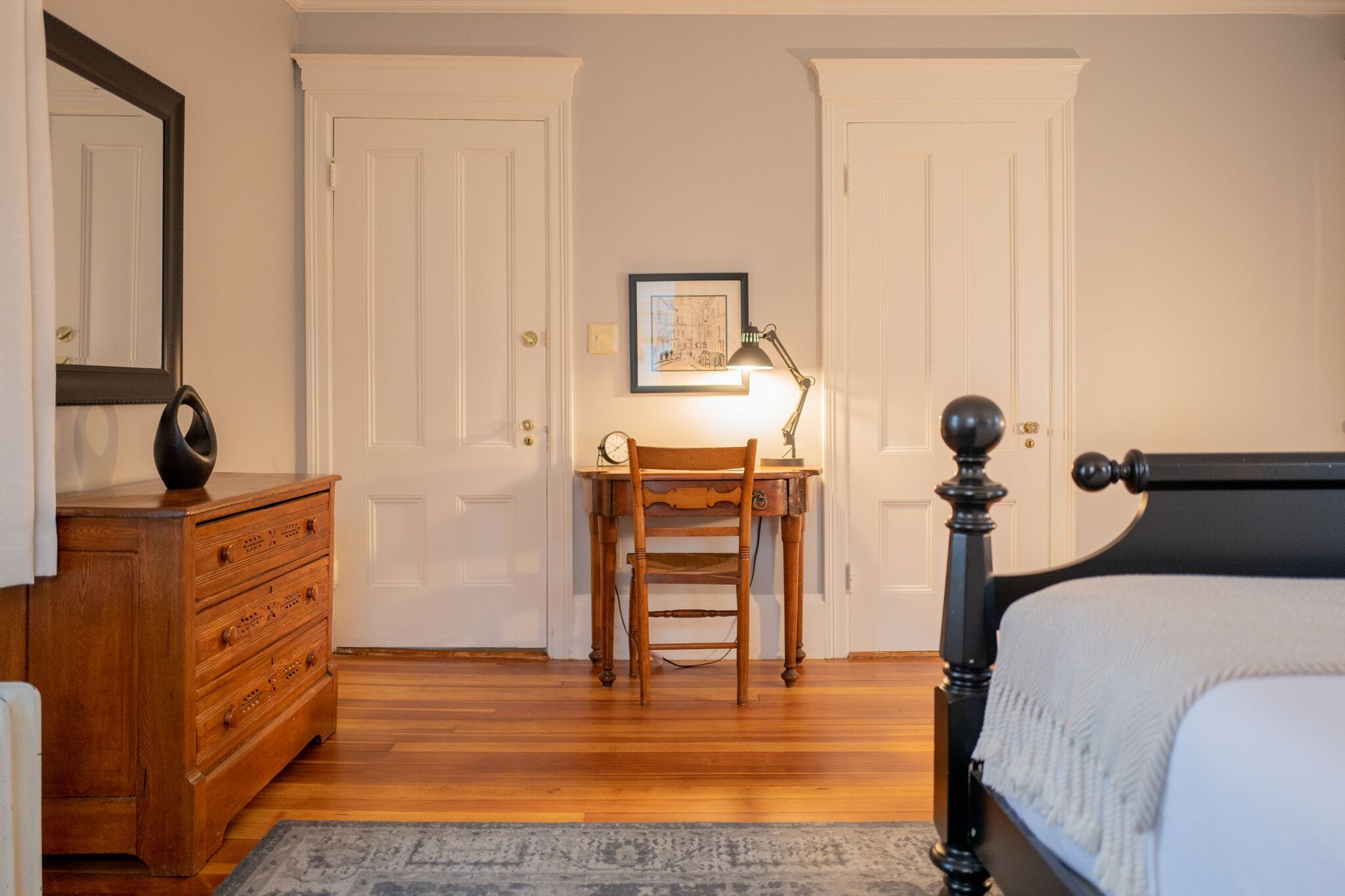 Une chambre à coucher avec une commode en bois, une chaise, un bureau avec une lampe et un lit doté d’une tête de lit noire.