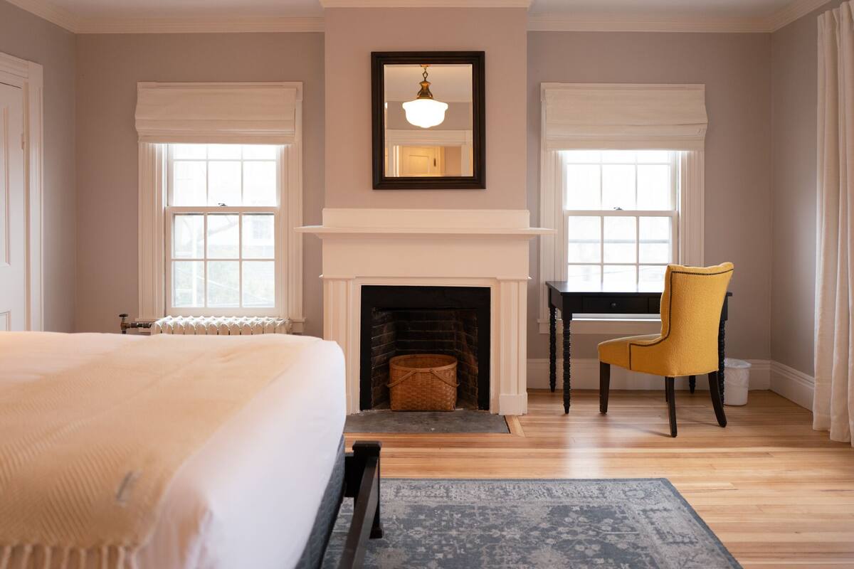 A bedroom with a large bed, a fireplace, a yellow armchair, and a view of the outside through two windows.