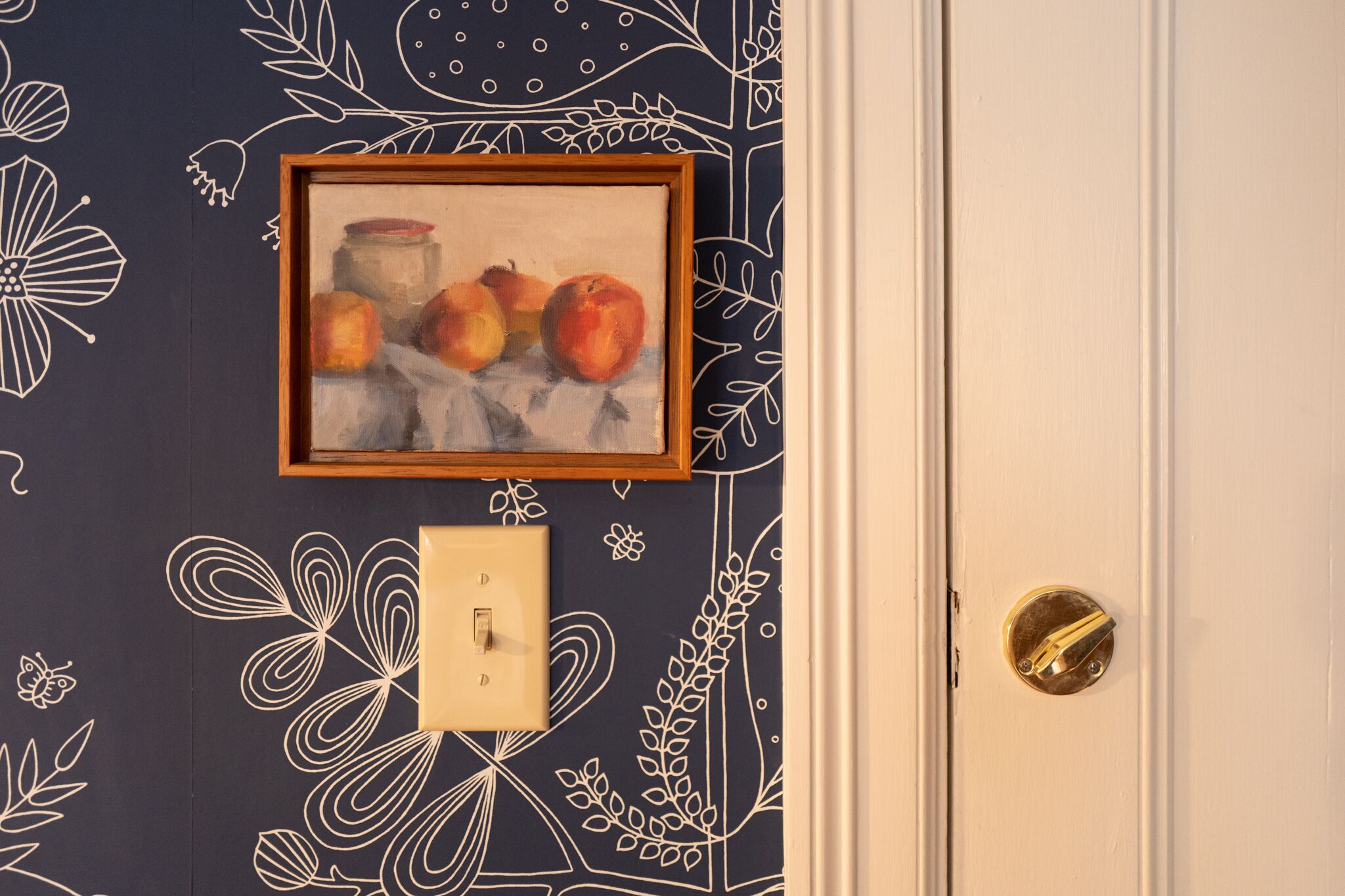 A door with a brass handle, a light switch, and a framed painting of fruit on the wall.