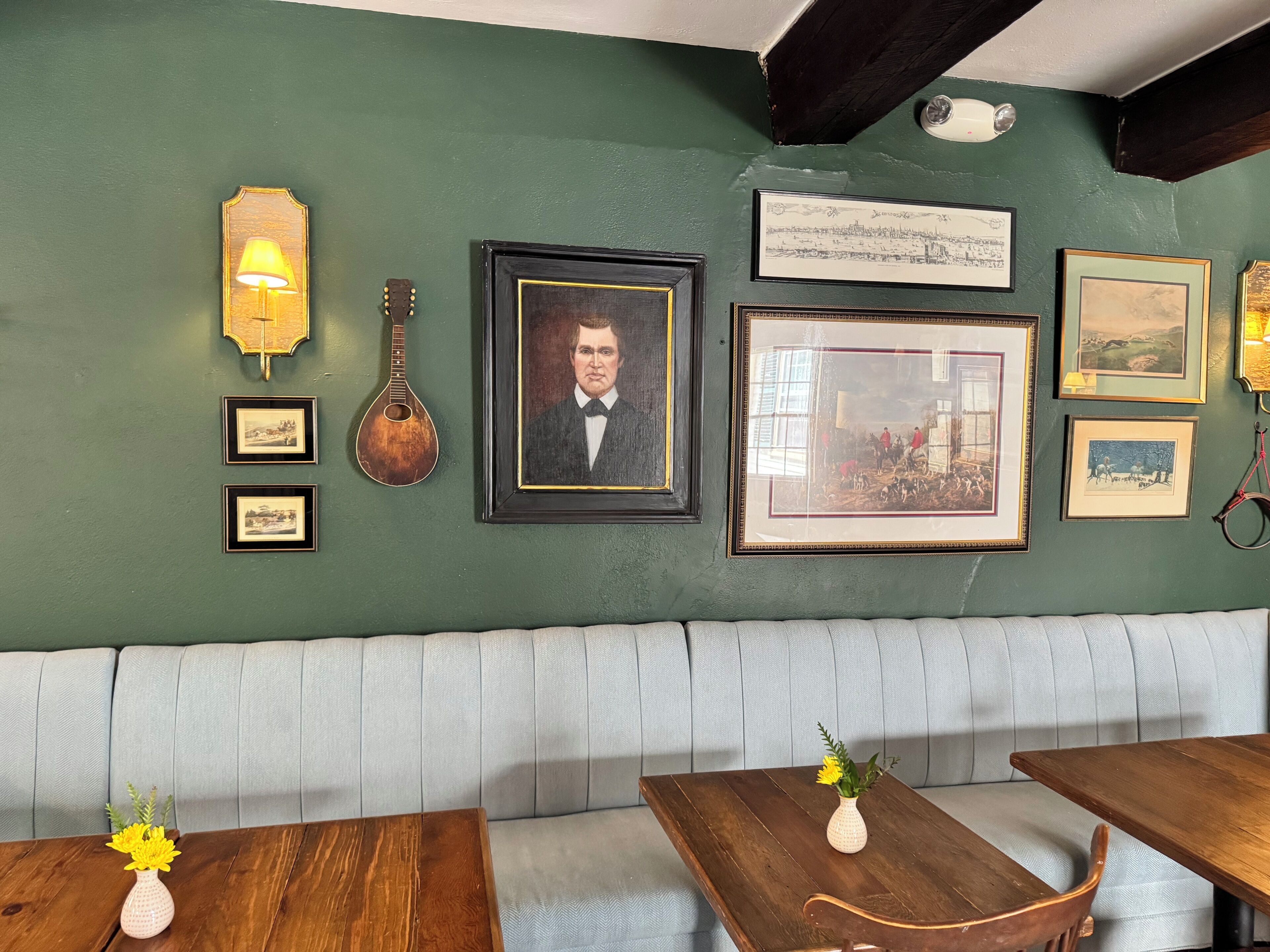 Una habitación con pared verde, una mesa de madera, un banco acolchado, varias imágenes enmarcadas y una guitarra.