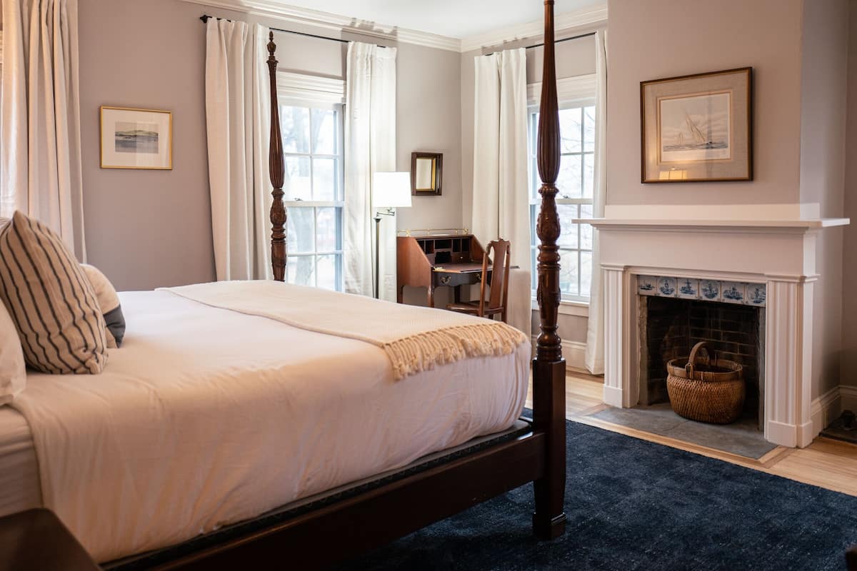 A four-poster bed with a dark wooden frame, a white bedspread, and striped pillows. There is a fireplace with a blue-tiled surround and a wicker basket on the floor.