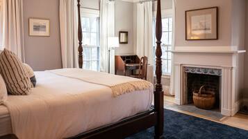 A four-poster bed with a dark wooden frame, a white bedspread, and striped pillows. There is a fireplace with a blue-tiled surround and a wicker basket on the floor.
