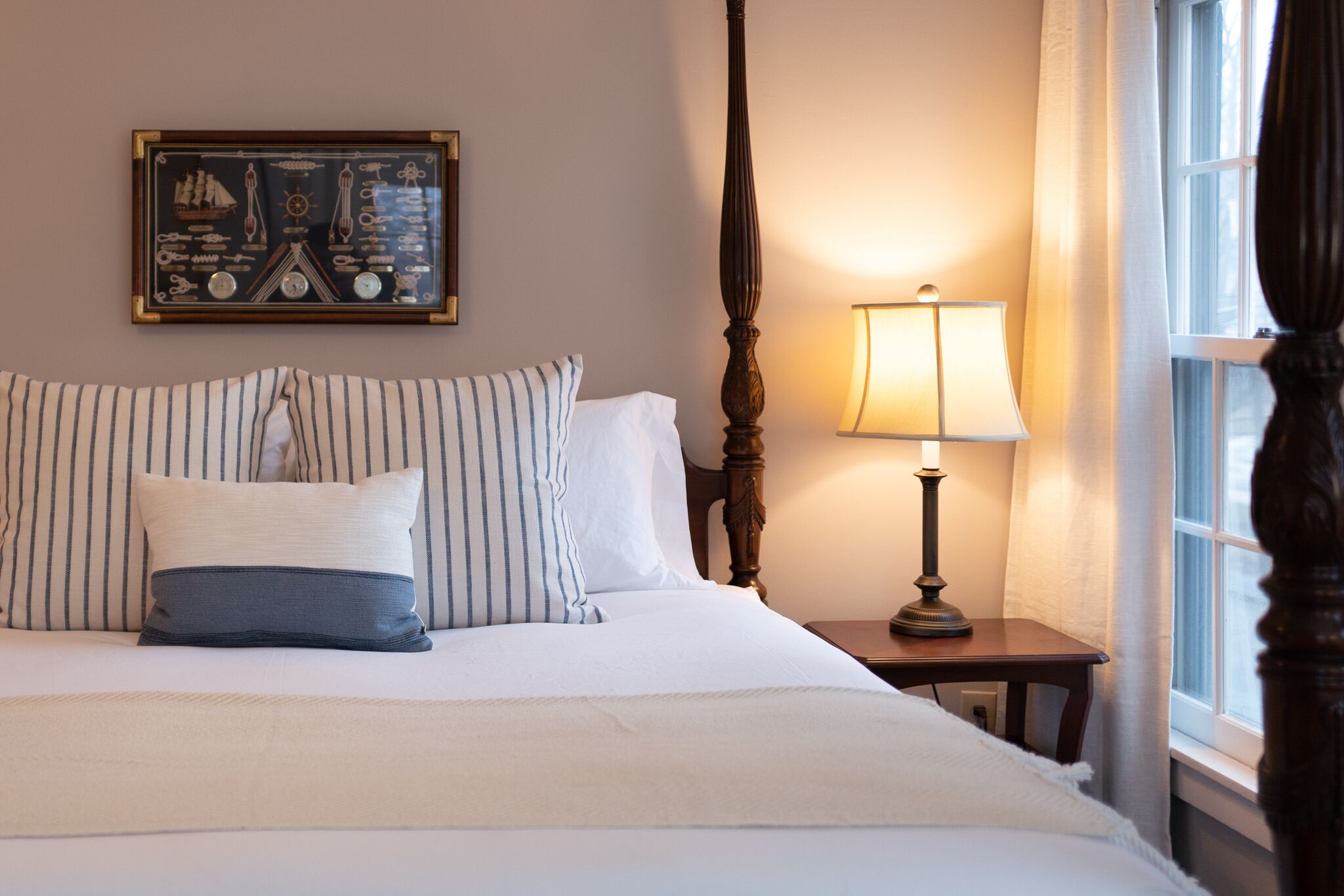 A neatly made bed with striped pillows, a framed nautical-themed artwork above it, a classic wooden bedside table with a lamp, and a window with curtains.