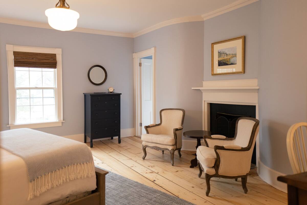 A room with a bed, a fireplace, two armchairs, a small table, and a dresser.
