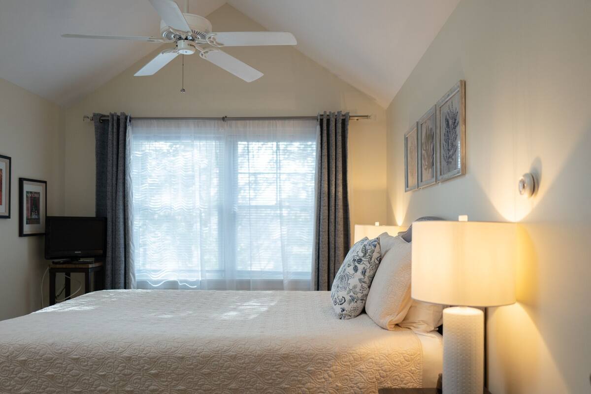 A hotel room with a bed, a ceiling fan, a TV, a lamp, and a window with curtains.