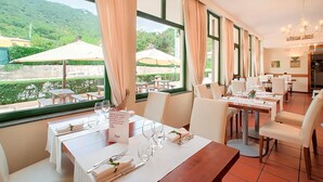 Un restaurant avec des tables dressées pour le service, de grandes fenêtres ornées de rideaux et une vue sur la verdure et les bâtiments environnants.
