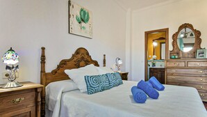 A neatly made bed with a patterned pillow, a wooden headboard, a nightstand with a lamp, a dresser with a mirror, and a painting on the wall.