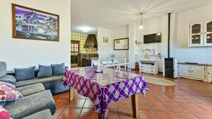 A living room with a sofa, a dining table with a purple tablecloth, a kitchen area with a stove, and a TV mounted on the wall.