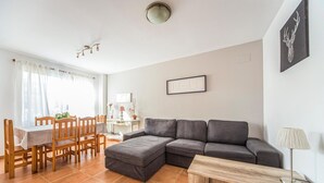 Ein Wohnzimmer mit grauem Ecksofa, einem Holzespeisetisch mit Stühlen, einem kleinen weißen Beistelltisch und einer Lampe.