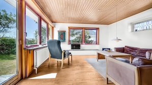 Ein modernes Wohnzimmer mit großen Fenstern, einer Holzbalkendecke, einem Sofa, einem Sessel und einem Esstisch.