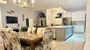 A modern kitchen with a central island, a dining area with floral-patterned chairs, and a flat-screen TV mounted on the wall. - Pool home in a quiet neighborhood (Cape Coral)