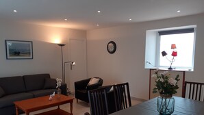 Ein Wohnzimmer mit Sofa, Esstisch, Stühlen, Couchtisch, Stehlampe, einer Uhr und einem Fenster mit Blick.