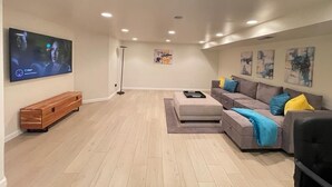 Uma sala de estar moderna com uma TV de tela plana, um banco de madeira, um sofá cinza com almofadas coloridas e arte abstrata na parede.