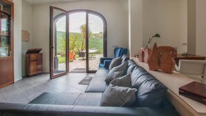 A modern living room with a leather sofa, a wooden cabinet, and a glass door leading to a patio with a view of a green landscape.