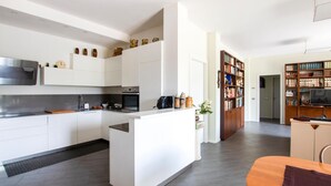 A modern kitchen with white cabinetry, a built-in oven, and a microwave. There is a small island with a sink and a coffee machine. The kitchen leads to a dining area with a wooden table and chairs.