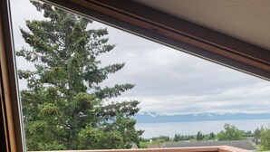 A wooden deck with a metal railing, a view of a parking lot with a red car, and a tree with green leaves. - VRBO unit in private home, in town with ocean views (Homer)
