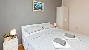 A neatly made bed with two pillows, a bedside lamp, a small bedside table with a plant, and a painting on the wall.