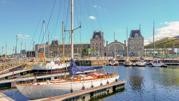 Ein Yachthafen mit mehreren dort liegenden Booten, ein großes Gebäude mit markantem Bogenportal und einem strahlend blauen Himmel.