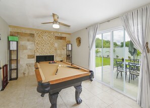 Una mesa de billar en una habitación con ventilador de techo, un atril para billar y vista a un jardín por medio de un ventanal.