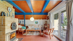 Un salon chaleureux avec une cheminée, des poutres apparentes en bois et une grande fenêtre donnant sur l’extérieur.