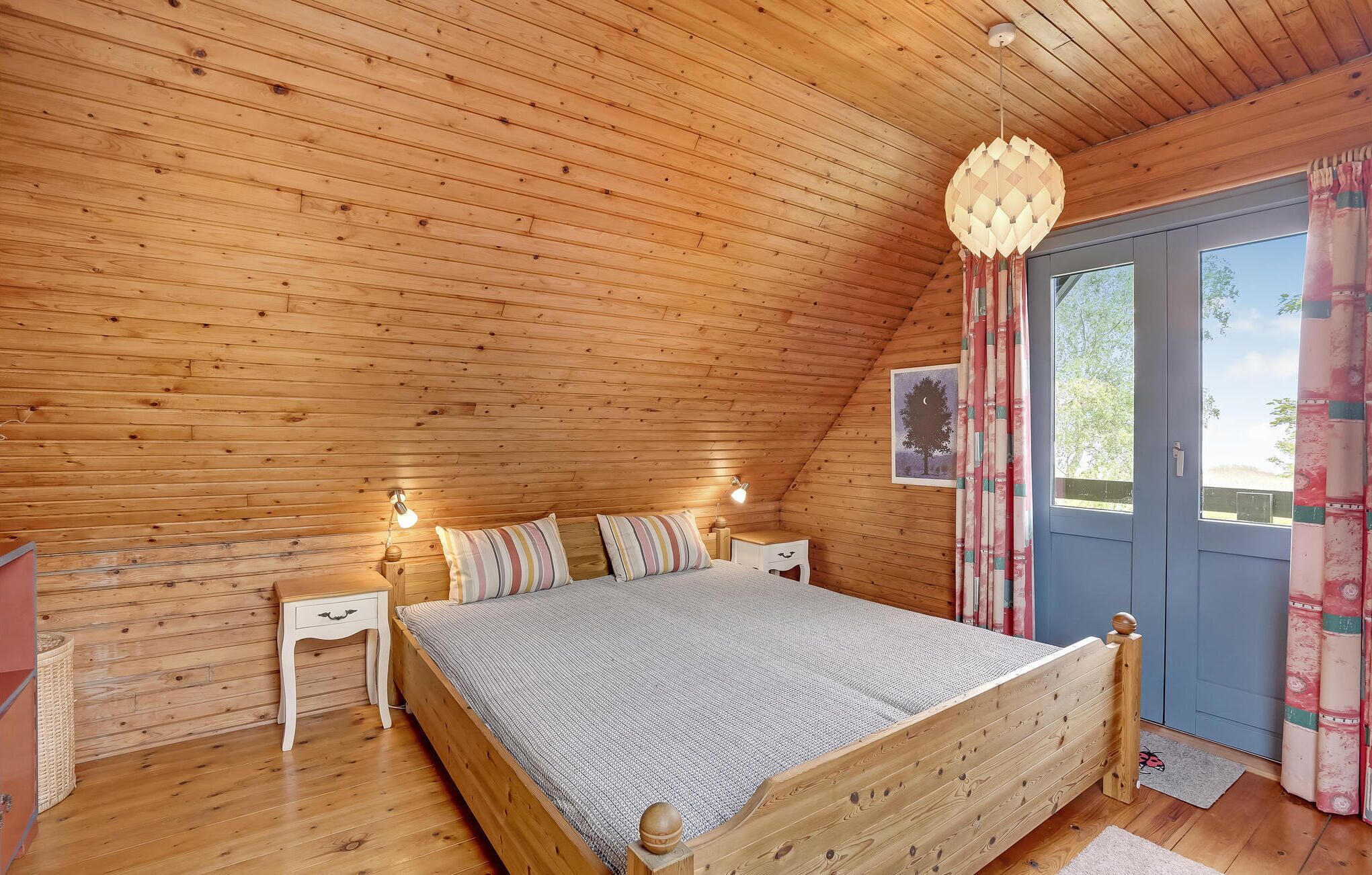 Une chambre confortable avec un plafond en bois, un lit agrémenté d’un coussin à rayures, des lampes de chevet et une fenêtre avec des rideaux.
