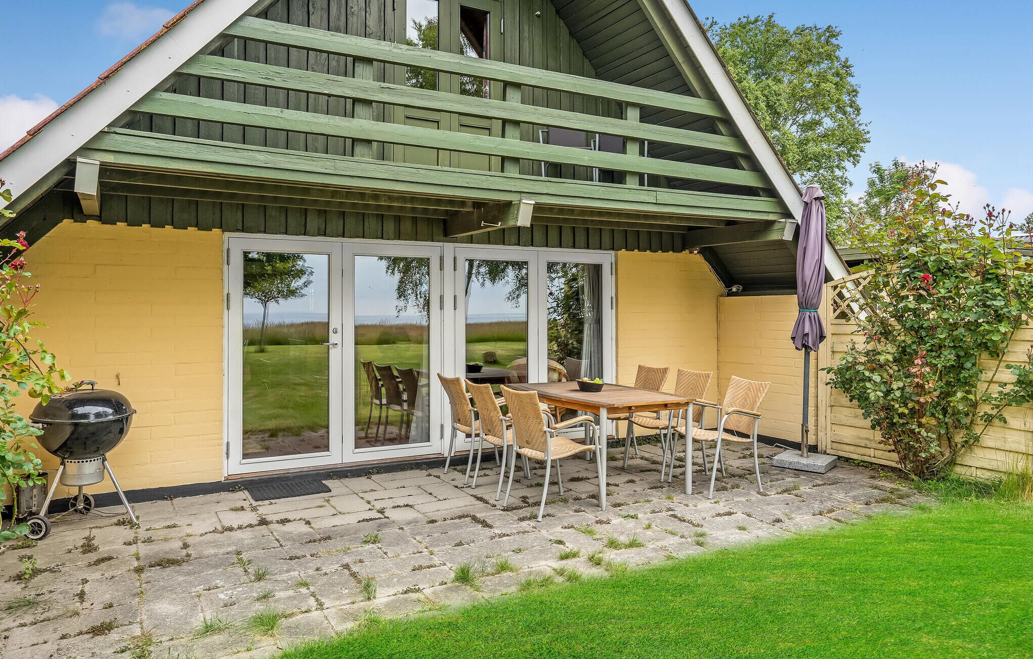 A covered patio with a table and chairs, a grill, and a view of the sea through glass doors.