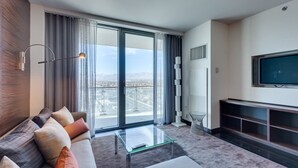 A modern living room with a sofa, a glass coffee table, a flat-screen TV, and a large sliding glass door leading to a balcony with a city view.