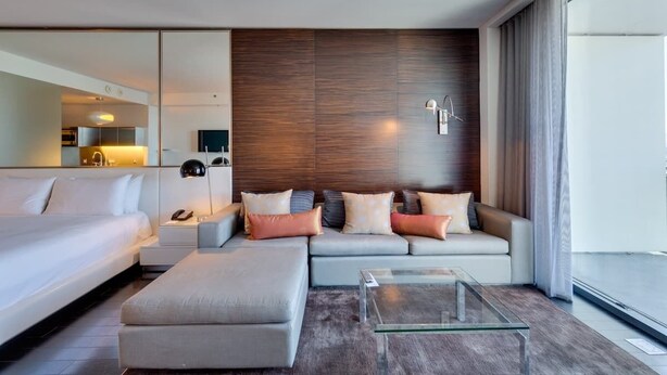 A modern hotel room with a large bed, a sofa, a glass coffee table, and a wooden accent wall.