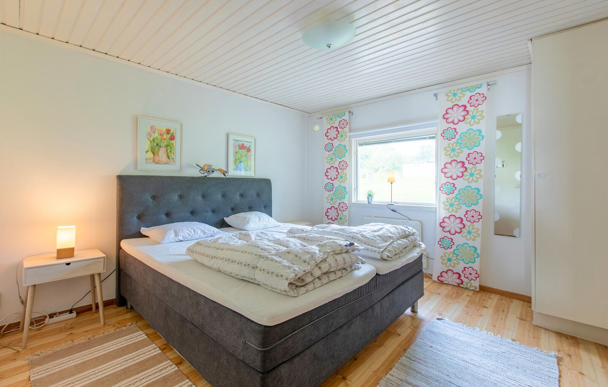 A bedroom with a grey upholstered bed, a bedside table with a lamp, and a window with floral curtains.