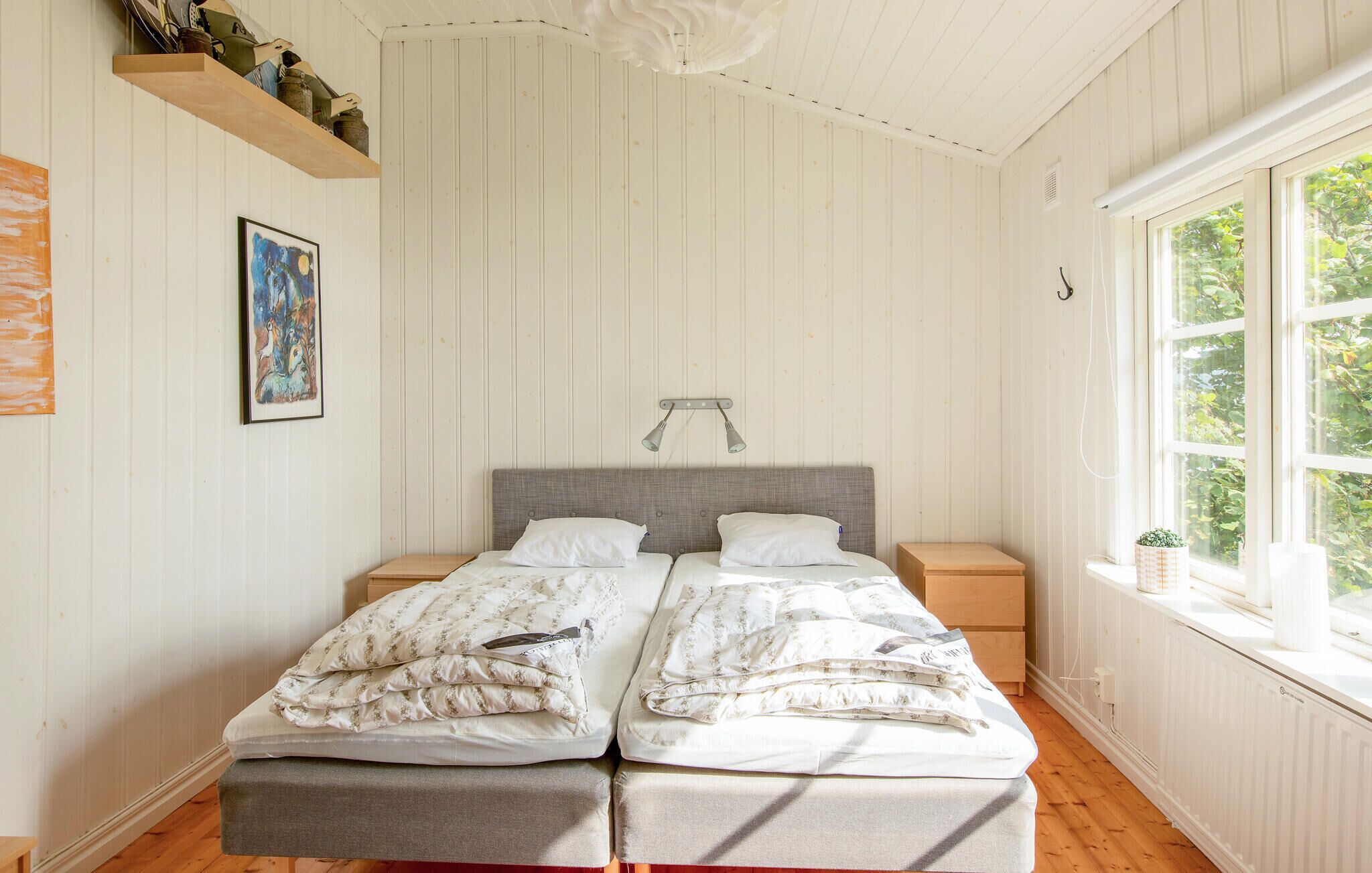 A bedroom with a double bed, two bedside tables, a headboard, a window with a view of greenery, and a wall-mounted light fixture.