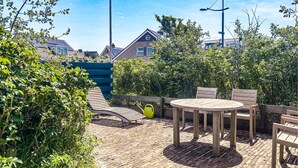 A patio area with a round wooden table and chairs, surrounded by greenery and a brick floor. - Beautiful (sta)caravan in Bergen aan Zee with WiFi (Bergen aan Zee)