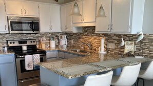 A modern kitchen with white cabinets, stainless steel appliances, a granite countertop, and a bar area with white stools. - Beautifully Remodeled Bay Front Condo (Navarre)