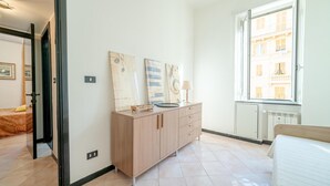 A hotel room with a wooden cabinet, a bed, a window with a view of buildings, and a door leading to another room.