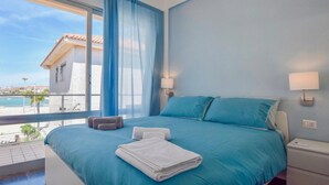 Quarto com cama, duas luminárias de cabeceira, janela com cortinas e vista para o mar e edifícios.