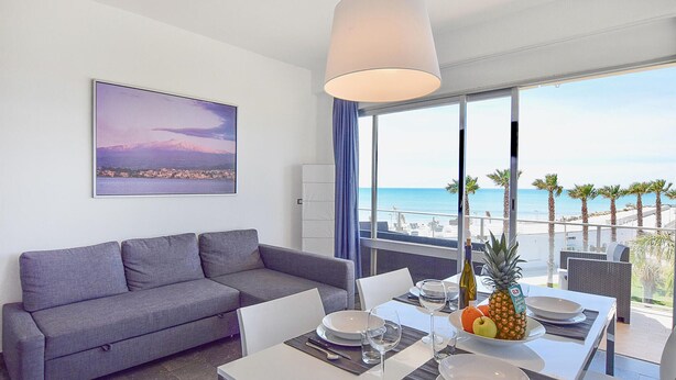 Uma sala de estar moderna com um sofá cinzento, uma mesa de jantar posta para dois e uma grande janela com vista para o mar e palmeiras.