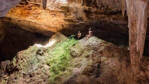 A cave with stalactites and stalagmites, two people sitting on a rock formation, and a light source illuminating the cave. - Cenote Pueblo Fantasma (Chemax)