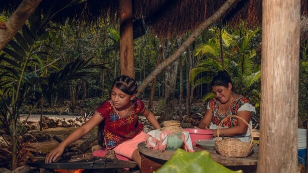 Two individuals cooking outdoors with a fire, surrounded by tropical vegetation. - Cenote Pueblo Fantasma (Chemax)