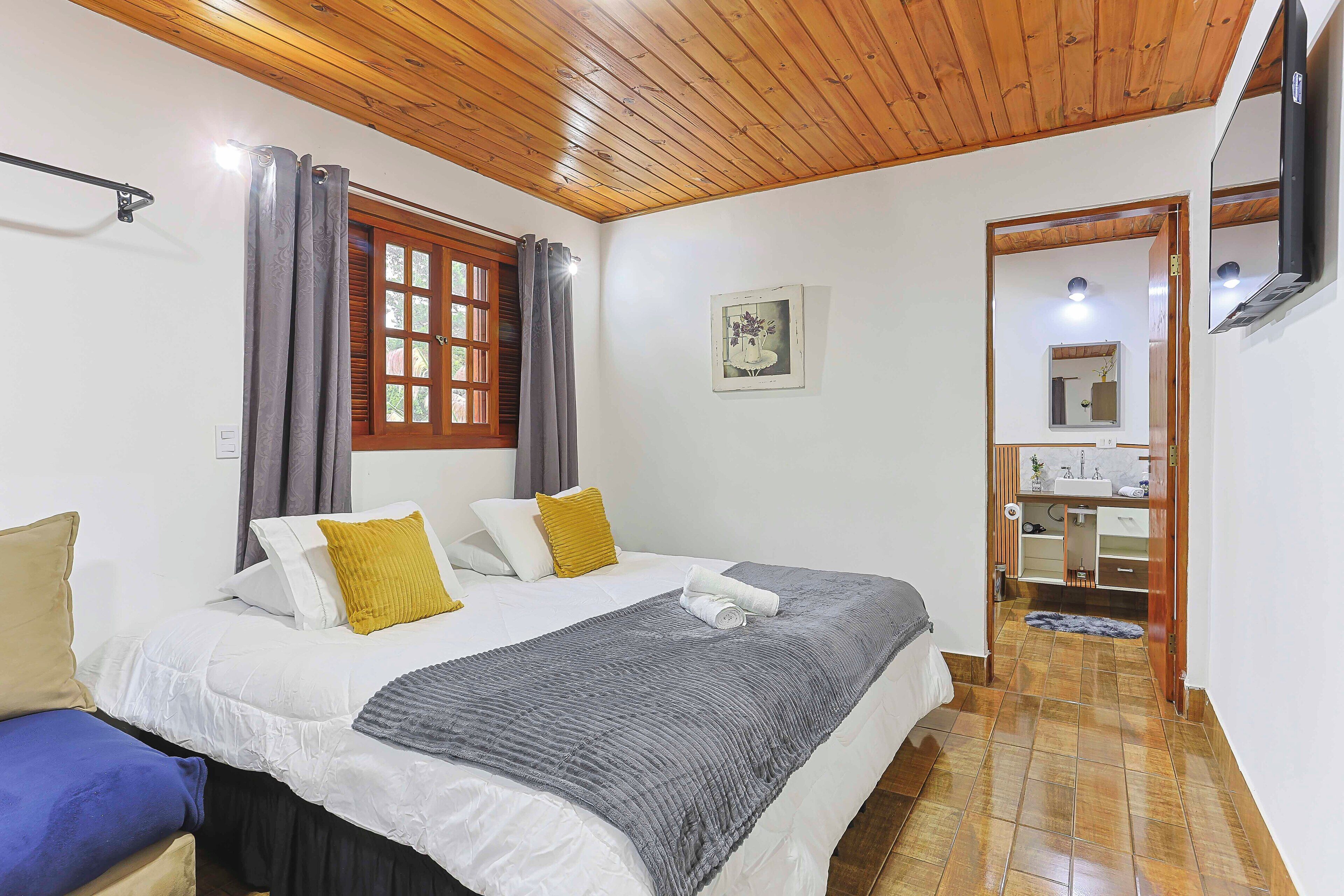 A bedroom with a bed, curtains, a window, a mirror, and a bathroom visible through an open door.