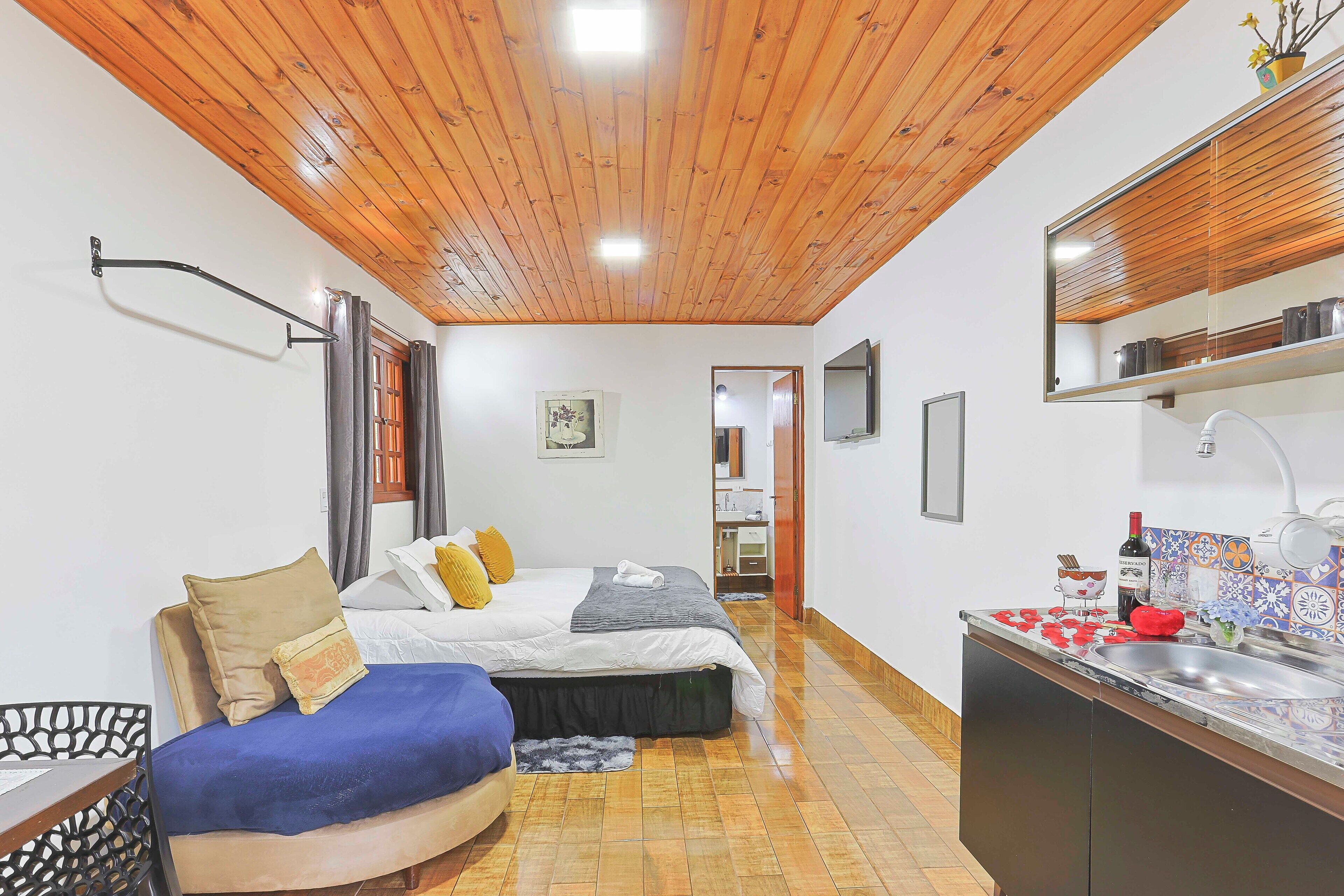 Habitación de hotel compacta con techo de madera, cama con almohadas, una pequeña zona de estar, kitchenette y baño visible a través de una puerta abierta.
