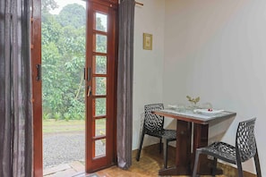 A room with a wooden door leading to a garden, a table with chairs, and a small table with a vase and glasses. - LoftS Campos 01 (Campos do Jordão)