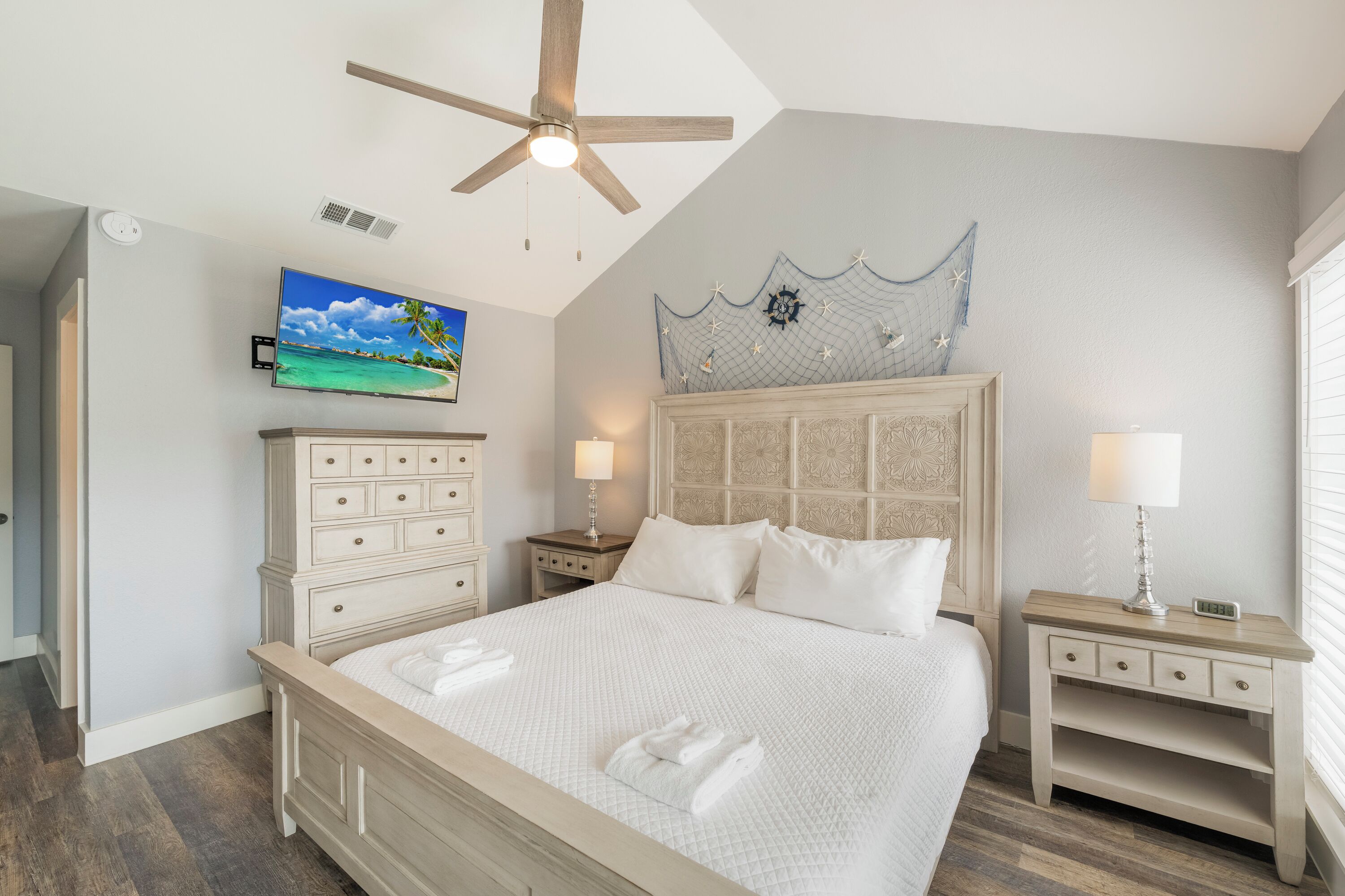 Une chambre à coucher comprenant un lit, des tables de chevet, un ventilateur de plafond, un téléviseur et un élément décoratif mural.