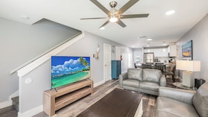 A modern living room with a flat-screen TV, leather sofas, a ceiling fan, and a kitchen in the background. - Poolside Condo | Modern Charm, Heated Pool & Steps to Beach (Corpus Christi)