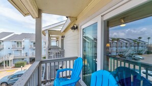 A balcony with blue chairs, a glass door, and a view of neighboring buildings. - Poolside Condo | Modern Charm, Heated Pool & Steps to Beach (Corpus Christi)