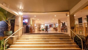Vestíbulo de hotel con una recepción, una escalera que sube a los pisos superiores y un área de descanso con mesas y sillas.
