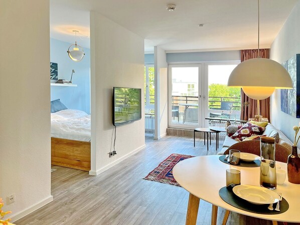 A modern hotel room with a bed, a TV mounted on the wall, a dining area with a table set for two, and a large window offering a view of the outdoors.