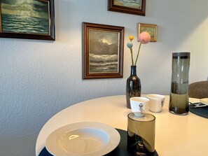 Uma mesa redonda com um prato branco, duas chávenas brancas com desenho de água-viva, um copo de vidro escuro e um recipiente de vidro alto.