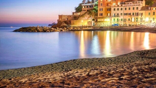 A coastal hotel with a castle-like building, a beach with a metal barrier, and calm waters reflecting the structure. - The traveler's choice (Genova)