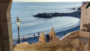 A view of a coastal area with a stone staircase leading to a beach, a pier, and a building with a balcony overlooking the sea. - The traveler's choice (Genova)