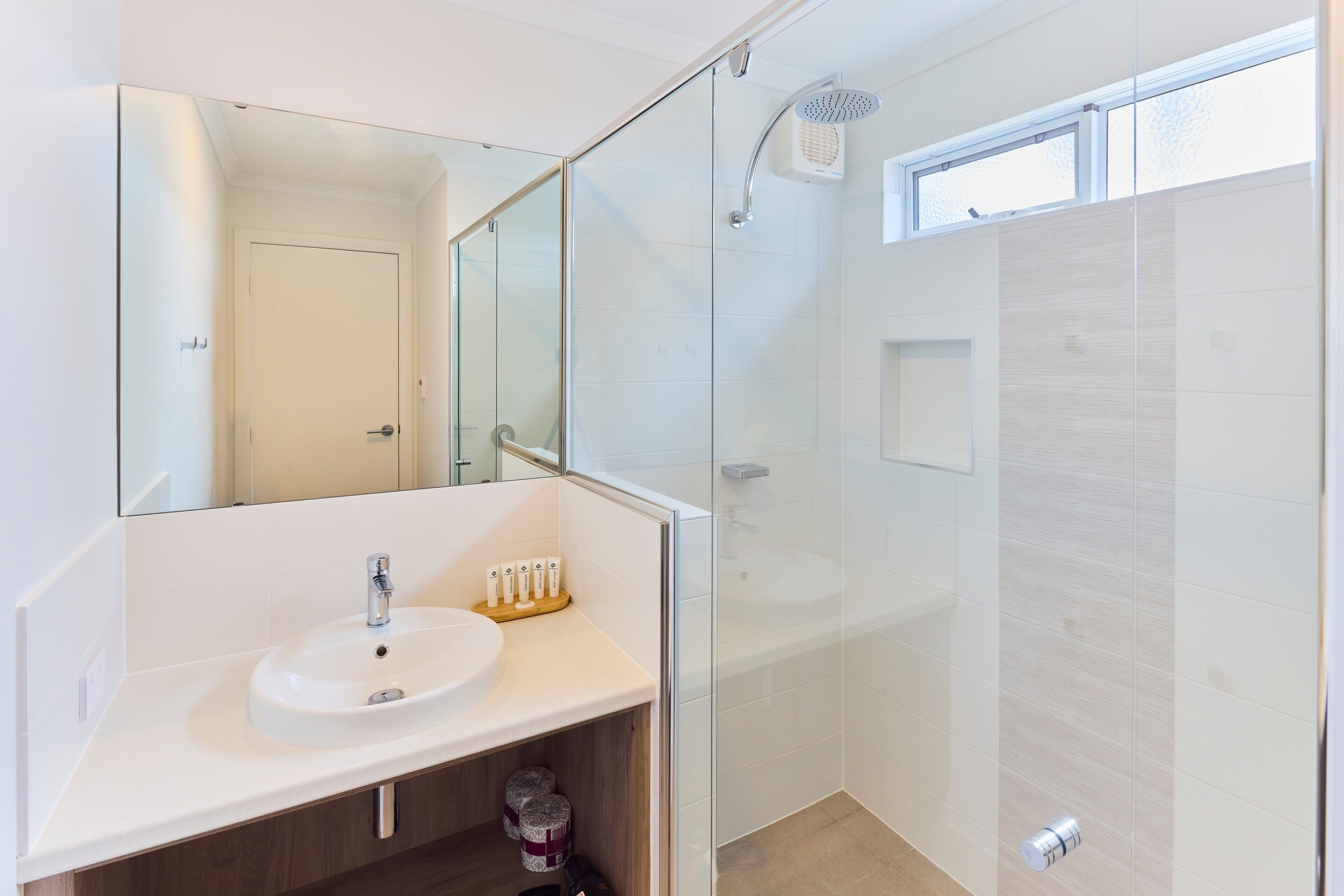 Un baño moderno con cabina de ducha de vidrio, un lavabo blanco con espejo encima y un ventanal sobre la ducha.
