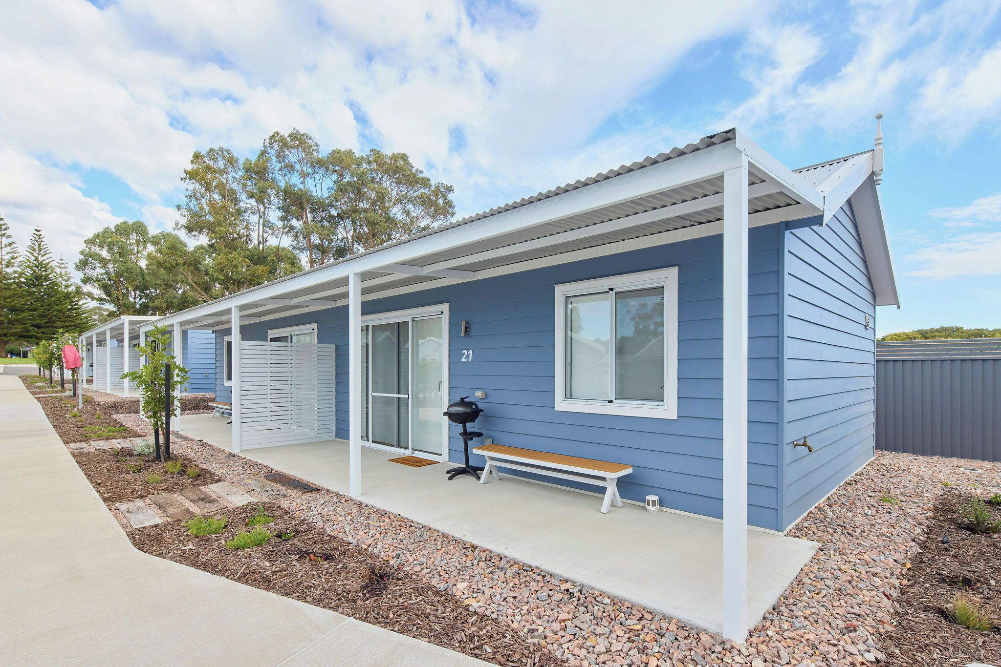 A row of blue mobile homes with white trim, numbered 21, featuring a covered porch area with a bench and a barbecue grill.